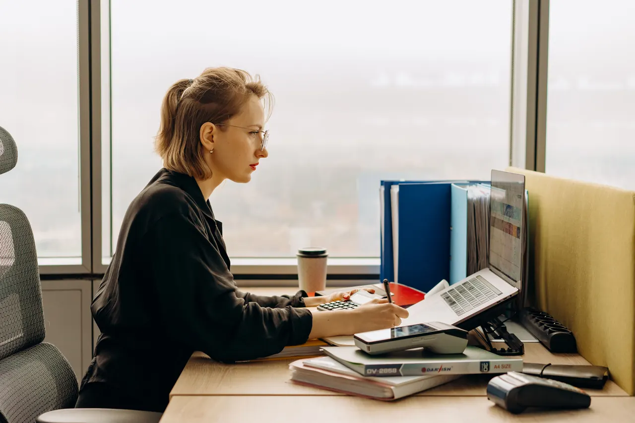 A woman working on a laptop with a calculator in a bright modern office